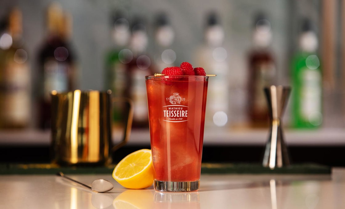 A clear glass contains a red drink topped with three raspberries on a gold pick. In the background there is half a lemon, and cocktail making equipment. Also in the background there are syrup bottles and spirit bottles.