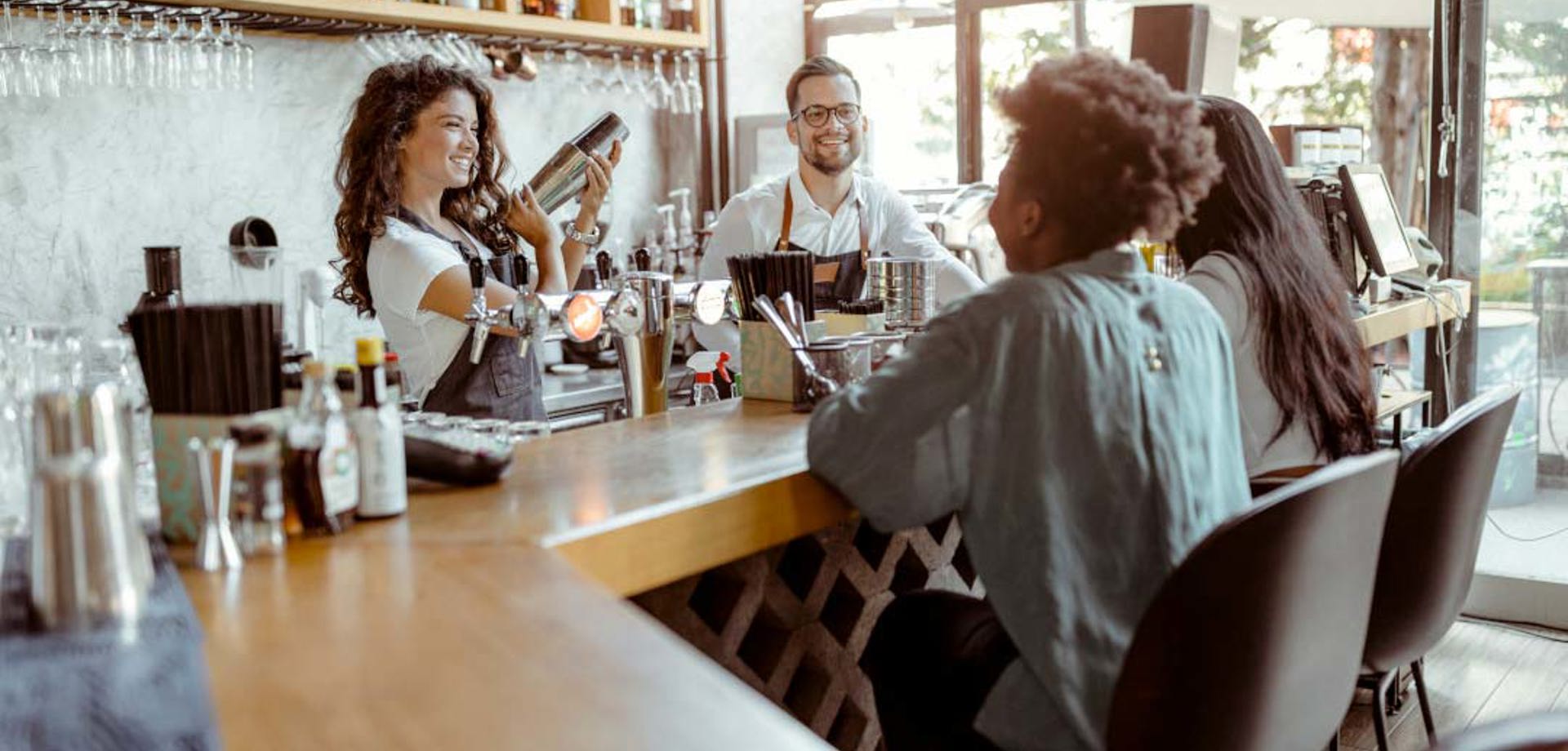 Two girls are sitting at the bar and talking to the workers in the cafe.