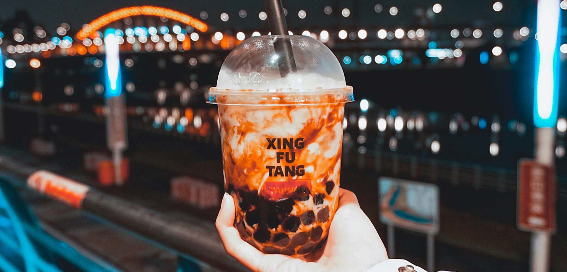 A hand holds a bubble tea in the air. In the background we can see a city at night.