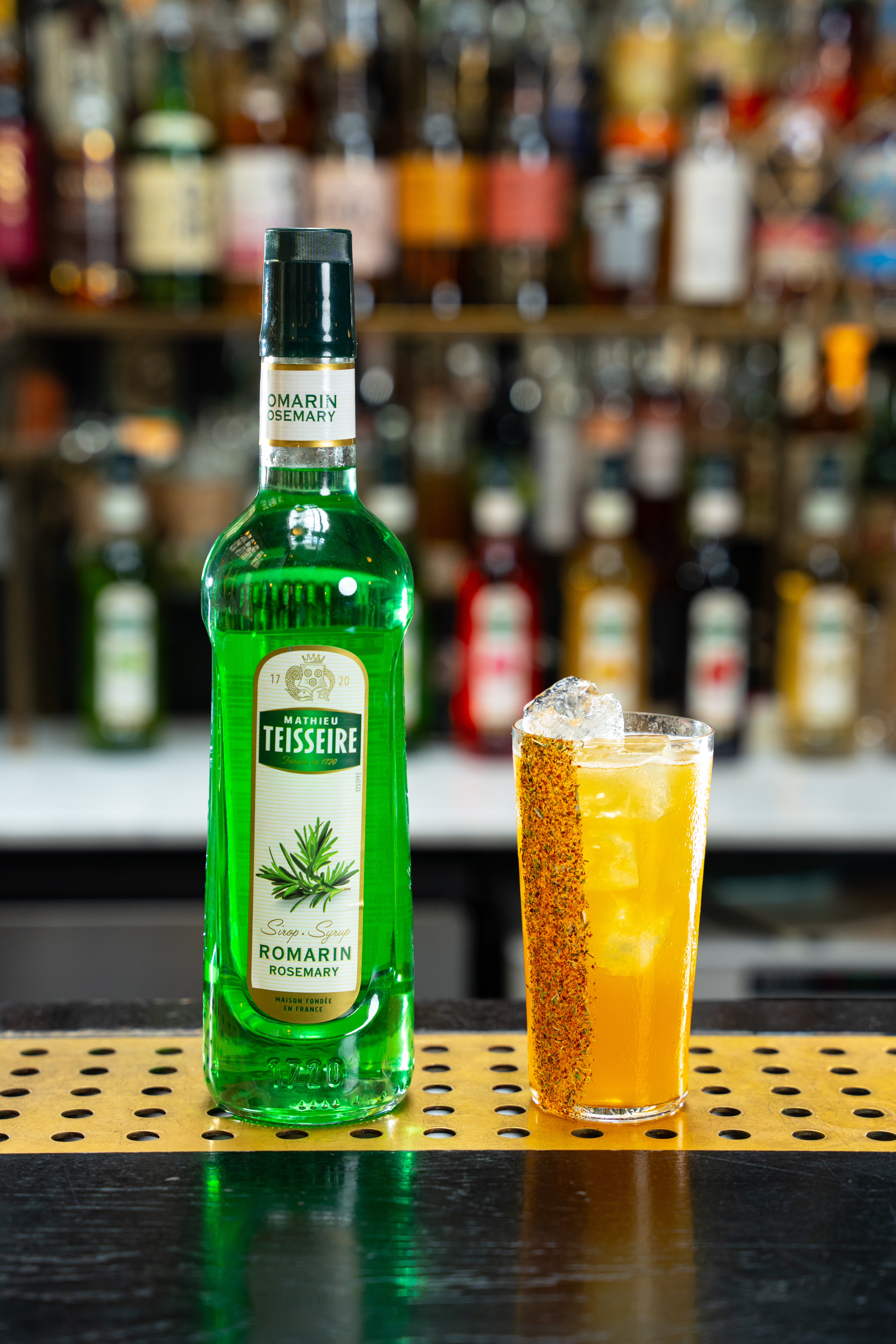 A tumbler glass is on a bar surface, placed next to a bottle of Mathieu Teisseire rosemary syrup. The glass contains a yellow drink, and the edge of the glass is lined with rosemary and hot pepper. 
