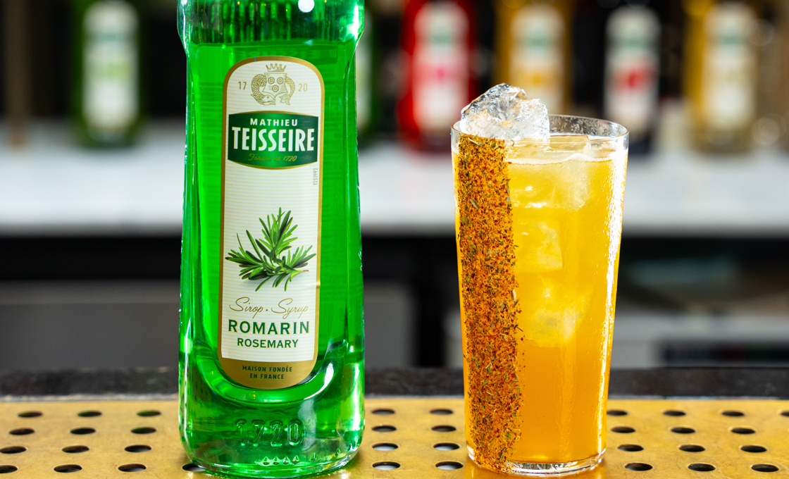 A tumbler glass is on a bar surface, placed next to a bottle of Mathieu Teisseire rosemary syrup. The glass contains a yellow drink, and the edge of the glass is lined with rosemary and hot pepper. 