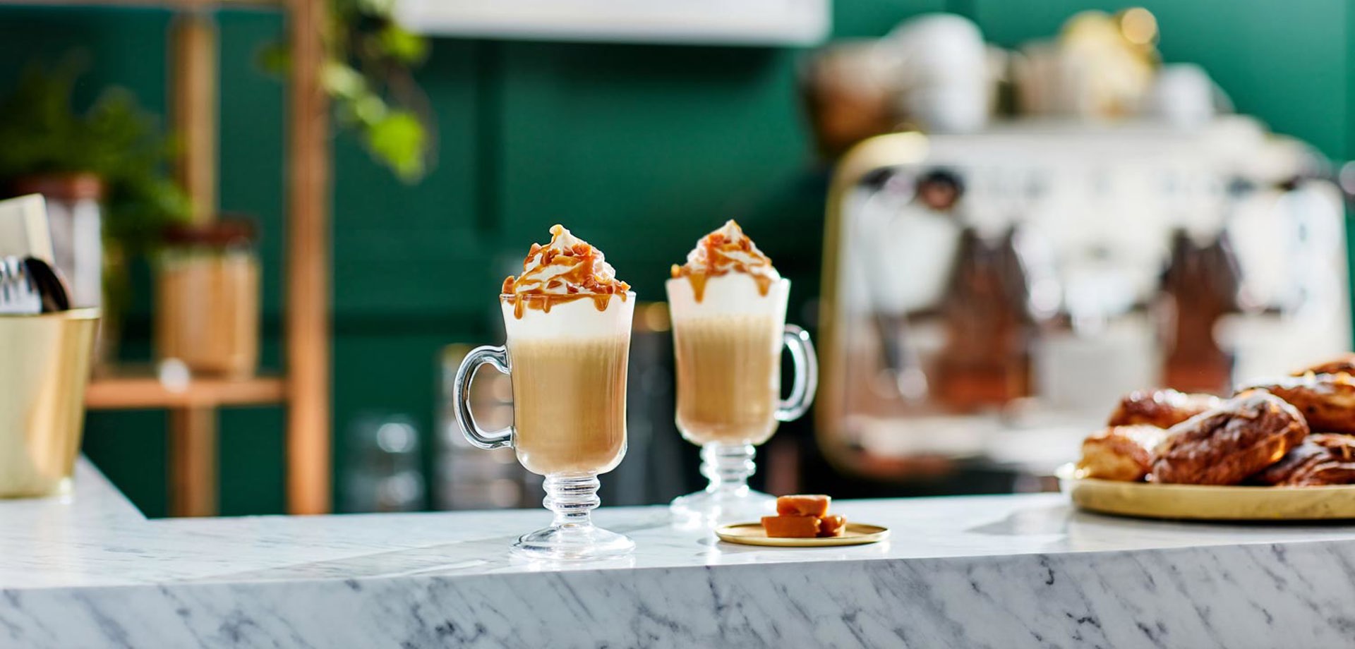 Two lattes sit atop a white marble surface. The lattes are topped with cream and caramel syrup. In the background there are pastries and a large coffee machine.