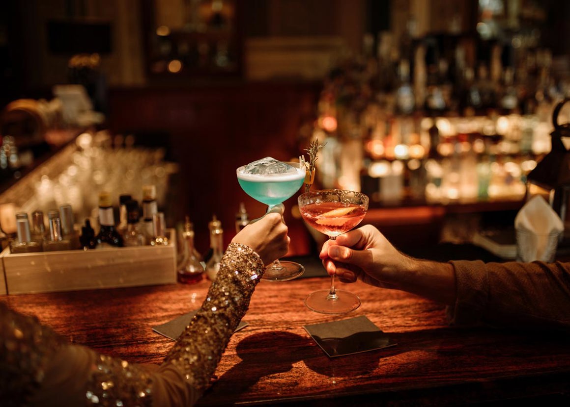 Two people are sat at a cocktail bar, we see them raising their glasses. The glasses contain a blue cocktail and a pink cocktail.