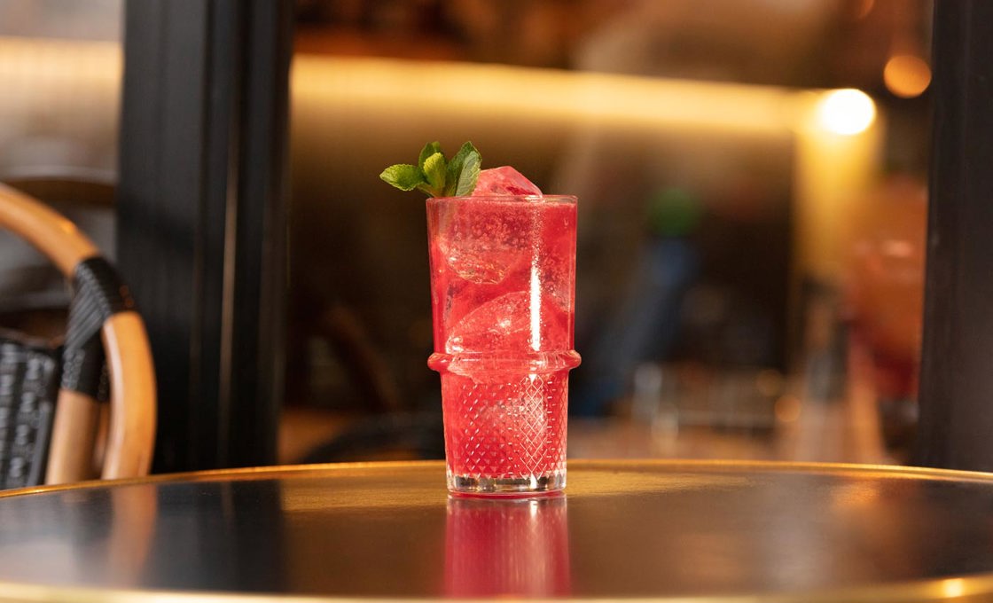 On an outdoor table in front of a large window sits a tall glass containing a red drink. The drink is topped with a mint leaf.