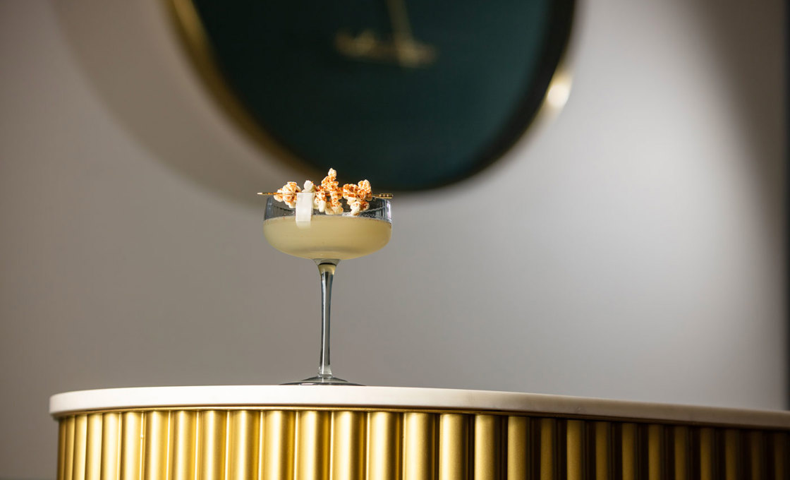 On a white table sits a short coupe glass filled with a margarita. Across the top of the glass is popcorn attached to a pick. In the background the room is dark, and we can see the edge of a green clock.