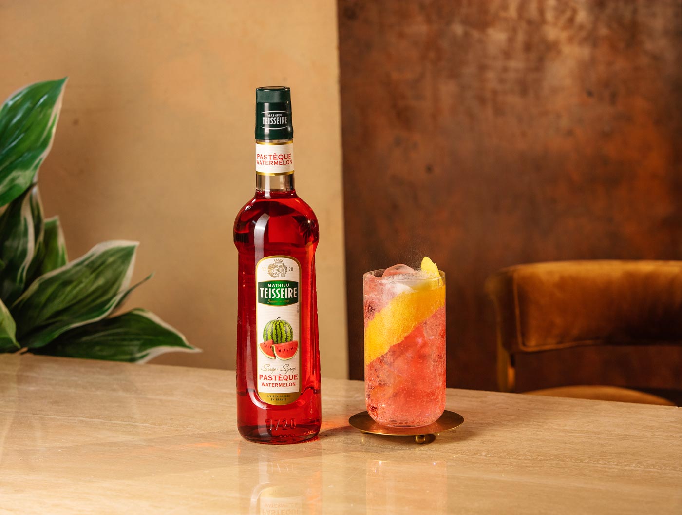 On a wooden table there is a bottle of watermelon Mathieu Teisseire syrup, and a tall glass containing ice, a pink drink and a slice of lemon. In the background there is a large plant and a leather bar chair.