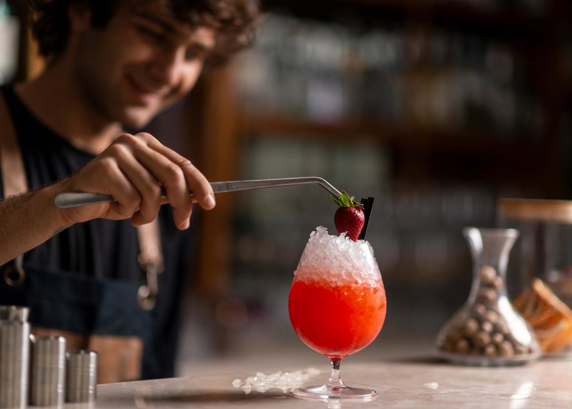 A man in an apron stands at a bar, using a long pair of tweasers to add a strawberry to the top of a drink. The drink is red and contains crushed ice. At the front of the bar there are different sized drinks measures.