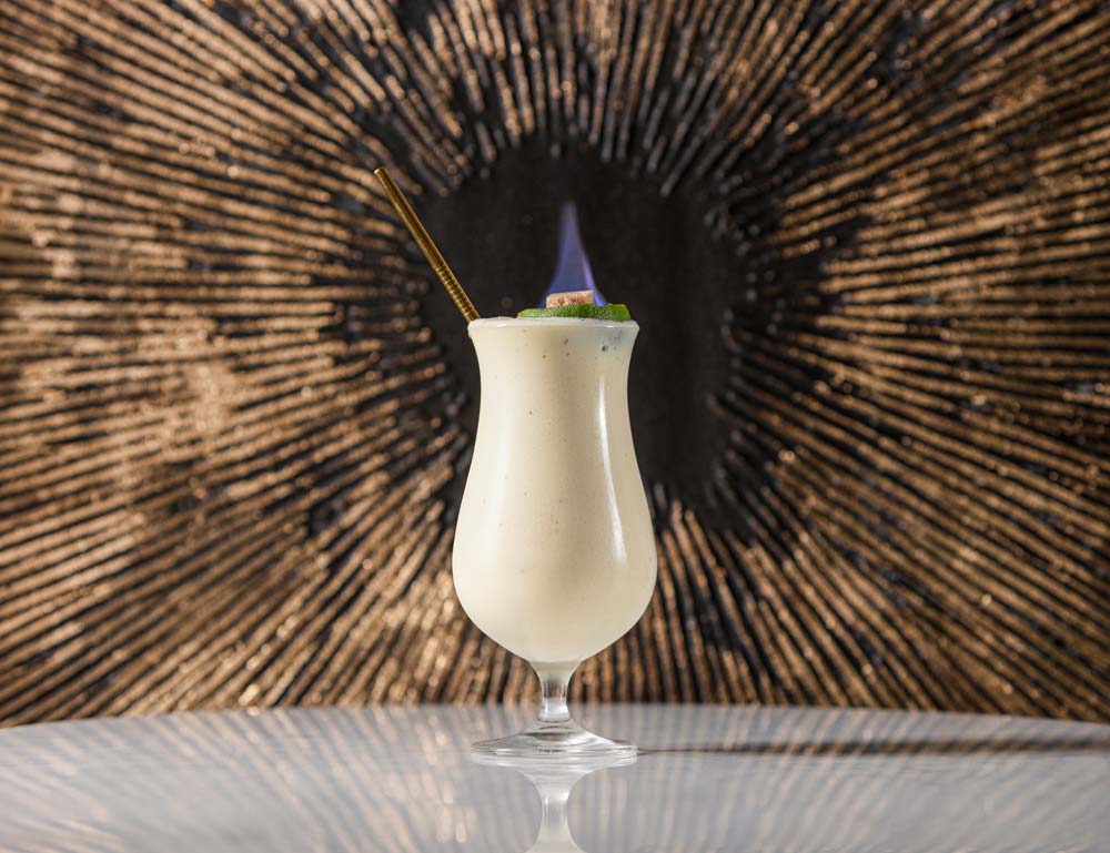 A hurricane glass is filled with a white drink. The drink is topped with a flaming sugar cube, and a gold straw is placed in the glass.
