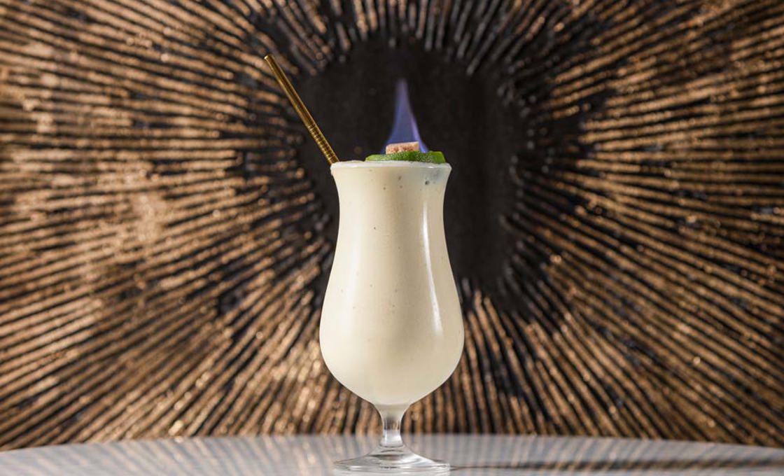 A hurricane glass is filled with a white drink. The drink is topped with a flaming sugar cube, and a gold straw is placed in the glass.