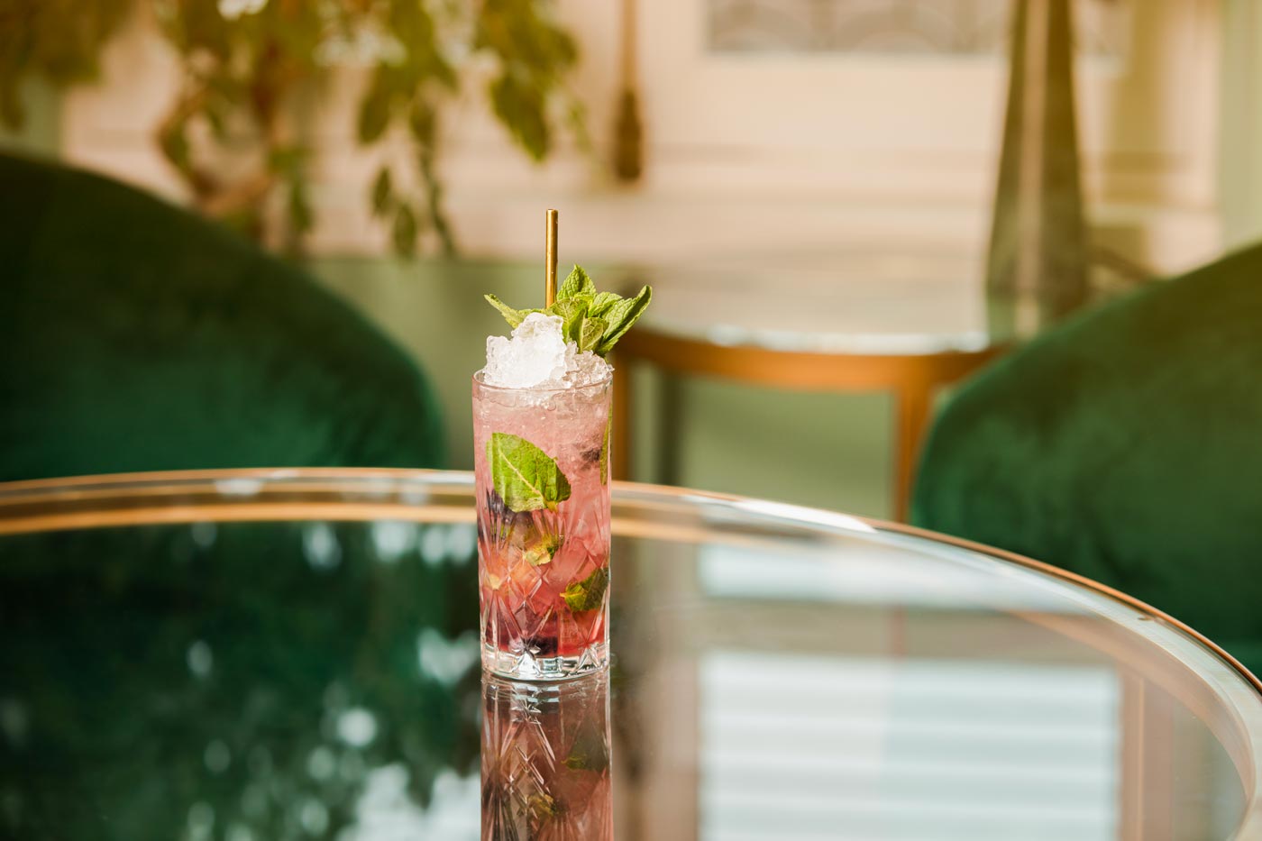 A tall glass is on a glass table. The glass contains a pink coloured drink and mint leaves. In the background we can see dark green velvet chairs and the egde of a large plant.