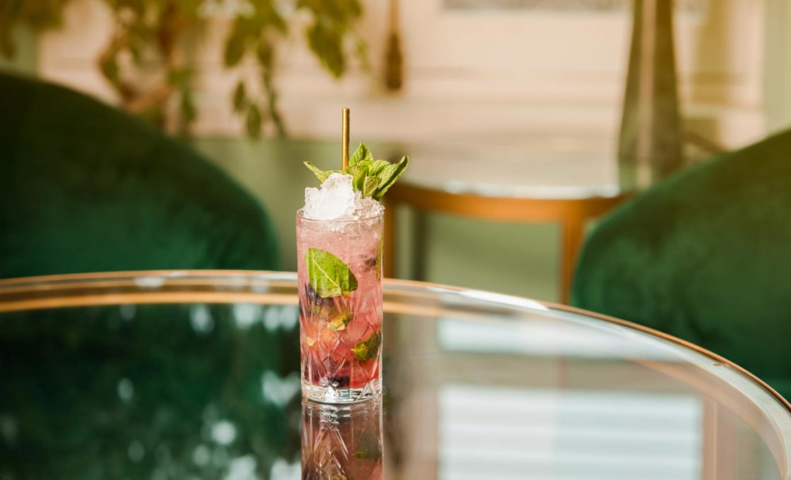 A tall glass is on a glass table. The glass contains a pink coloured drink and mint leaves. In the background we can see dark green velvet chairs and the egde of a large plant.