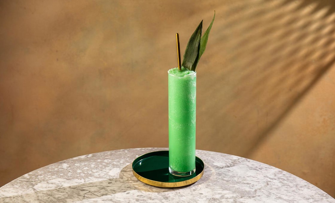 An ultra highball glass is placed on a large green coaster, which rests on a marble table. The glass contains an icy green liquid, a golden straw and a garnish of pineapple leaves.